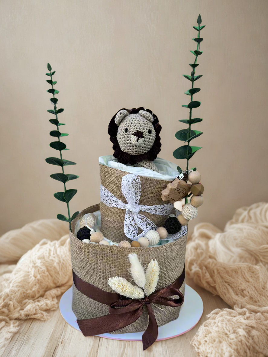 Two-tier neutral safari diaper cake featuring rolled white diapers wrapped in natural burlap and finished with delicate white lace and deep brown satin ribbon bows. The base tier is accented with dried floral stems for a soft rustic touch.

The top tier is crowned with a handmade crocheted lion plush in cream with a dark brown mane, peeking sweetly from the diapers. A coordinating wooden and silicone beaded pacifier clip in natural, black, and beige tones drapes along the side, complemented by a wooden anim