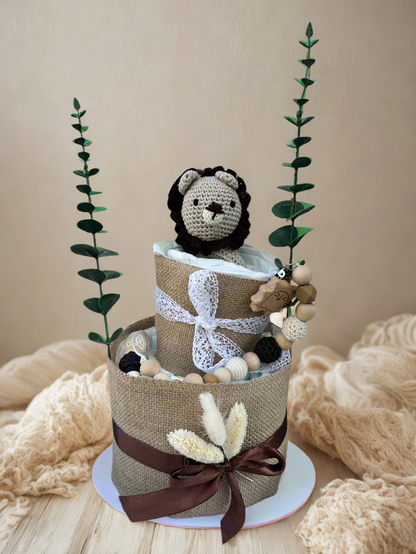Two-tier neutral safari diaper cake featuring rolled white diapers wrapped in natural burlap and finished with delicate white lace and deep brown satin ribbon bows. The base tier is accented with dried floral stems for a soft rustic touch.

The top tier is crowned with a handmade crocheted lion plush in cream with a dark brown mane, peeking sweetly from the diapers. A coordinating wooden and silicone beaded pacifier clip in natural, black, and beige tones drapes along the side, complemented by a wooden anim