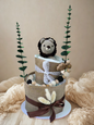 Two-tier neutral safari diaper cake featuring rolled white diapers wrapped in natural burlap and finished with delicate white lace and deep brown satin ribbon bows. The base tier is accented with dried floral stems for a soft rustic touch.

The top tier is crowned with a handmade crocheted lion plush in cream with a dark brown mane, peeking sweetly from the diapers. A coordinating wooden and silicone beaded pacifier clip in natural, black, and beige tones drapes along the side, complemented by a wooden anim