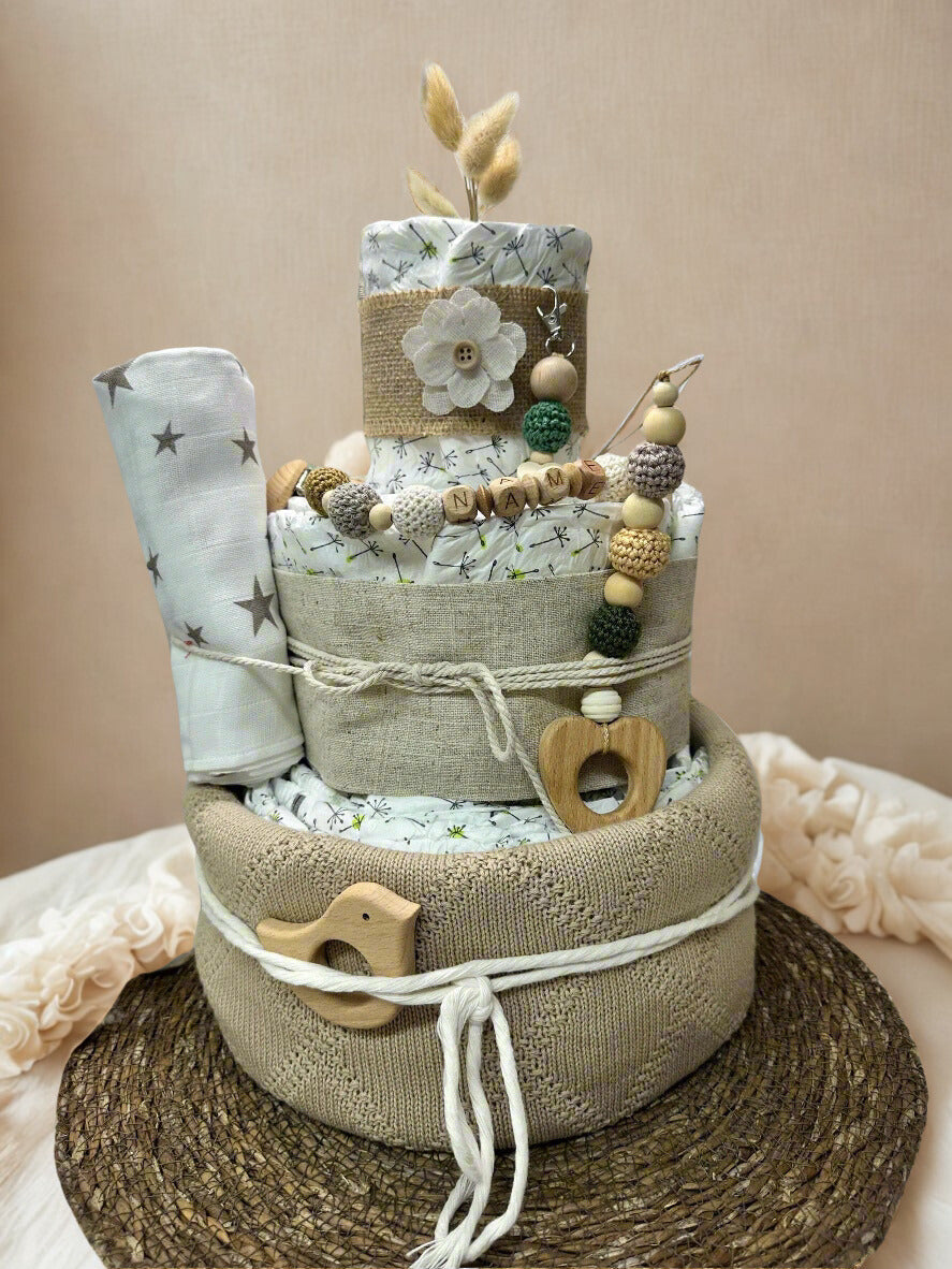 Neutral personalized diaper cake baby shower centerpiece featuring three tiers of rolled white diapers wrapped in soft sage and natural linen fabrics. The base tier is covered in a cozy knit-textured taupe fabric and finished with a rustic white cord, accented by a charming wooden bird teether for a sweet, minimalist touch.

The middle tier is styled with light linen wrap and coordinating rope detail, complemented by a silicone and crochet beaded pacifier clip in earthy tones of cream, moss green, mustard, 