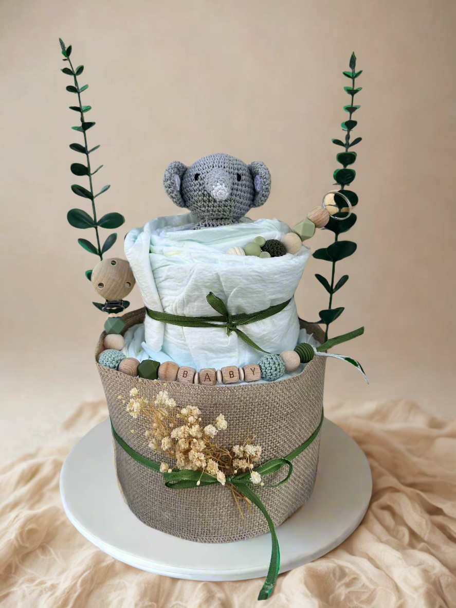 Two-tier green safari diaper cake featuring rolled white diapers wrapped in natural burlap and tied with deep green satin ribbon bows. A small crocheted gray elephant plush peeks out from the top tier, accented with soft green and wooden beaded details.

The base is decorated with a personalized wooden and silicone pacifier clip spelling “BABY” in sage and natural tones, along with coordinating teething beads and a wooden clip. Dried florals and tall faux greenery stems frame the arrangement, creating a mod