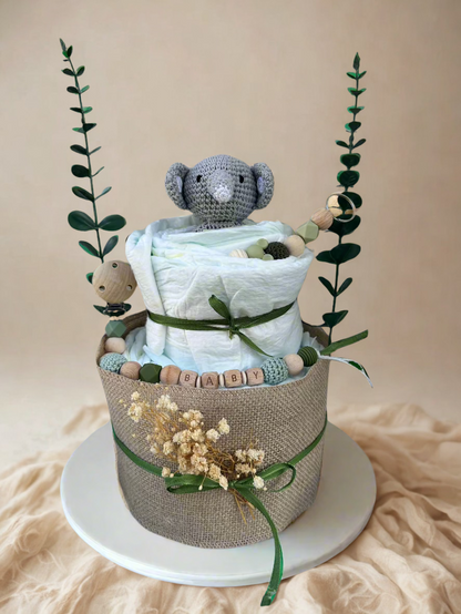 Two-tier green safari diaper cake featuring rolled white diapers wrapped in natural burlap and tied with deep green satin ribbon bows. A small crocheted gray elephant plush peeks out from the top tier, accented with soft green and wooden beaded details.

The base is decorated with a personalized wooden and silicone pacifier clip spelling “BABY” in sage and natural tones, along with coordinating teething beads and a wooden clip. Dried florals and tall faux greenery stems frame the arrangement, creating a mod