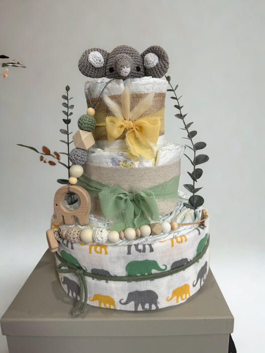 Safari elephant diaper cake baby shower centerpiece featuring three tiers of rolled white diapers wrapped in burlap ribbon and soft sage green fabric bows. Top tier decorated with a crocheted gray elephant plush, accented with dried bunny tail grass and eucalyptus greenery stems. Adorned with wooden elephant-shaped teething ring, silicone and crochet beaded pacifier clip in green, cream, and natural wood tones. Base tier wrapped in white fabric with green, yellow, and gray elephant print, creating a playful