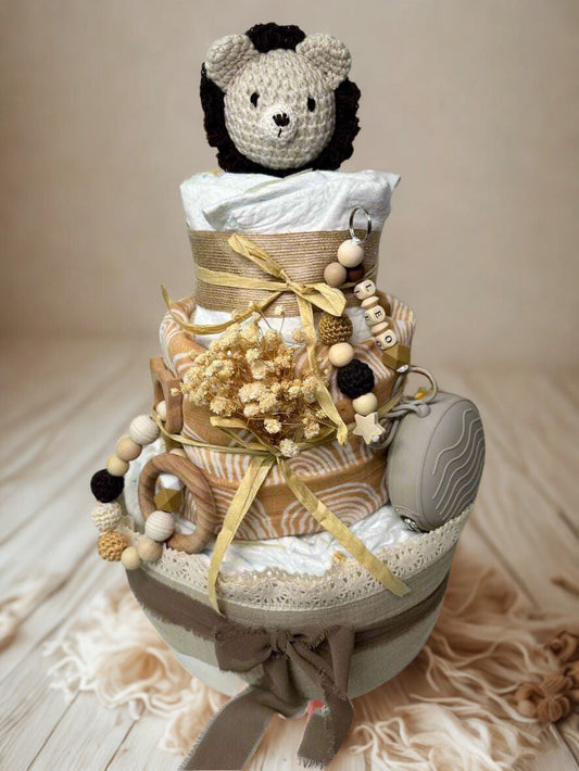Safari lion diaper cake baby shower centerpiece featuring three tiers of rolled white diapers styled in warm caramel, beige, and soft neutral tones. The base tier is wrapped in layered taupe and cream fabrics with delicate lace trim, finished with a soft brown ribbon bow for an elegant rustic foundation.

The middle tier showcases a modern mustard and cream rainbow-patterned fabric, accented with dried florals and tied with raffia ribbon for a natural boho touch. A coordinating wooden ring teether and silic