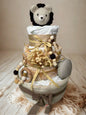 Safari lion diaper cake baby shower centerpiece featuring three tiers of rolled white diapers styled in warm caramel, beige, and soft neutral tones. The base tier is wrapped in layered taupe and cream fabrics with delicate lace trim, finished with a soft brown ribbon bow for an elegant rustic foundation.

The middle tier showcases a modern mustard and cream rainbow-patterned fabric, accented with dried florals and tied with raffia ribbon for a natural boho touch. A coordinating wooden ring teether and silic