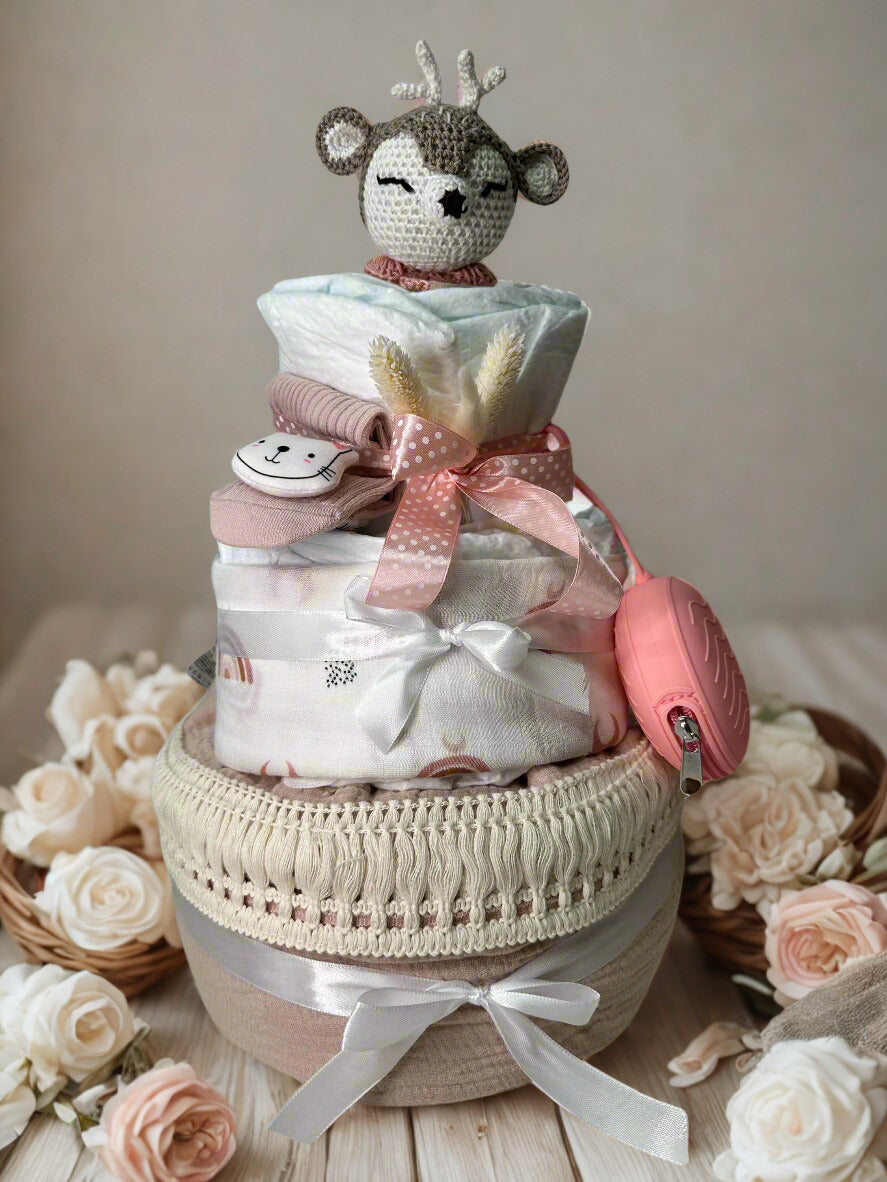 Blush woodland deer diaper cake baby shower centerpiece featuring three tiers of rolled white diapers styled in soft pink, ivory, and warm neutral tones. The base tier is wrapped in natural linen fabric and finished with delicate ivory macramé trim and a classic white satin ribbon bow for an elegant, vintage-inspired touch.

The middle tier is dressed in subtle rainbow-print fabric and tied with a crisp white bow, complemented by a soft pink pacifier case attached at the side for both beauty and practicalit
