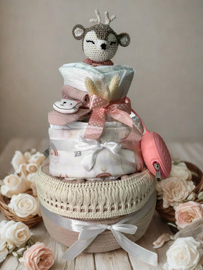 Blush woodland deer diaper cake baby shower centerpiece featuring three tiers of rolled white diapers styled in soft pink, ivory, and warm neutral tones. The base tier is wrapped in natural linen fabric and finished with delicate ivory macramé trim and a classic white satin ribbon bow for an elegant, vintage-inspired touch.

The middle tier is dressed in subtle rainbow-print fabric and tied with a crisp white bow, complemented by a soft pink pacifier case attached at the side for both beauty and practicalit