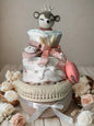 Blush woodland deer diaper cake baby shower centerpiece featuring three tiers of rolled white diapers styled in soft pink, ivory, and warm neutral tones. The base tier is wrapped in natural linen fabric and finished with delicate ivory macramé trim and a classic white satin ribbon bow for an elegant, vintage-inspired touch.

The middle tier is dressed in subtle rainbow-print fabric and tied with a crisp white bow, complemented by a soft pink pacifier case attached at the side for both beauty and practicalit