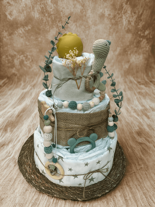 Three-tier dinosaur diaper cake baby shower centerpiece featuring rolled white diapers wrapped in soft sage fabric and rustic burlap ribbon. Top tier styled with a mustard yellow silicone pacifier case and delicate dried baby’s breath tied with natural twine. Accented with eucalyptus greenery stems for a fresh botanical touch. Middle tier decorated with a crocheted green dinosaur plush attached to a wooden teething ring, along with a coordinating silicone and crochet beaded pacifier clip in green, cream, an