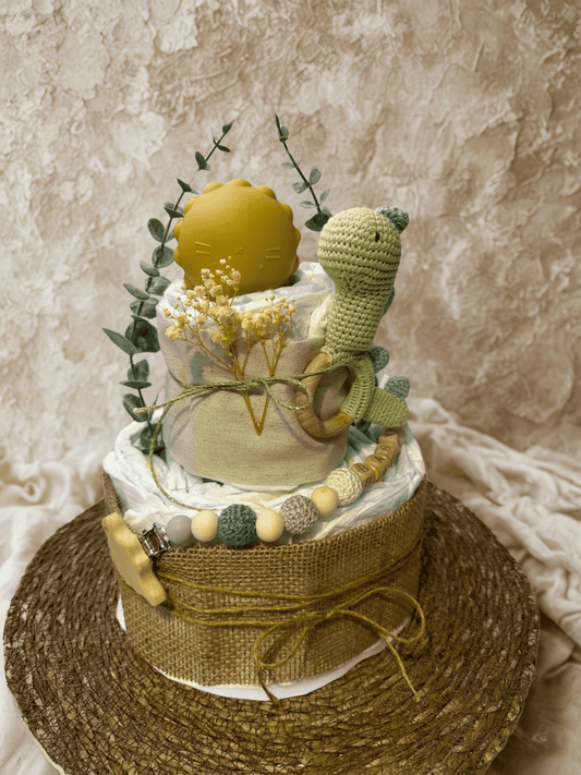 Dinosaur-themed diaper cake baby shower centerpiece featuring two tiers of rolled white diapers wrapped in soft sage green fabric and rustic burlap ribbon. Top tier decorated with a yellow silicone pacifier case and accented with delicate dried baby’s breath and eucalyptus greenery. Adorned with a crocheted green dinosaur plush attached to a natural wooden teething ring, plus a coordinating silicone and crochet beaded pacifier clip in green, cream, and gray tones with wooden letter beads spelling “BABY.” Ba