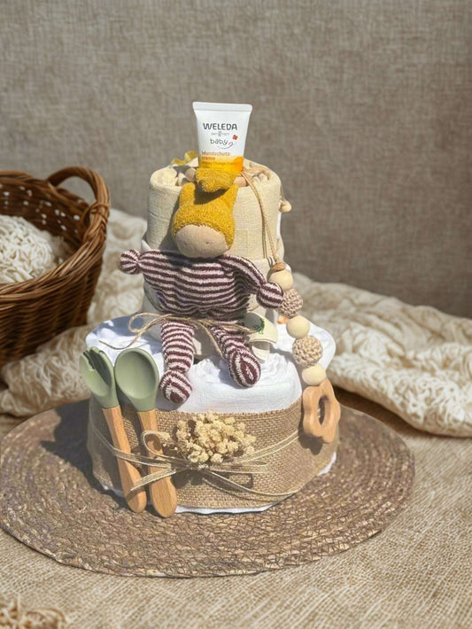 Neutral boho diaper cake baby shower centerpiece featuring two tiers of rolled white diapers wrapped in natural linen and rustic burlap ribbon. Top tier includes a tube of baby care cream and a soft yellow plush comfort toy, accented with a coordinating wooden and crochet beaded pacifier clip in warm beige tones.

Bottom tier decorated with dried florals and twine bow detail, complemented by sage green baby utensils with wooden handles and a natural wooden teething ring. Displayed on a woven rattan mat with