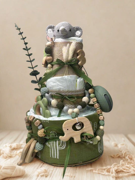 Green cactus-themed diaper cake baby shower centerpiece featuring three tiers of rolled white diapers wrapped in sage and olive green fabric with burlap trim and satin ribbon bows. Top tier decorated with a crocheted gray elephant plush, accented with dried wheat grass and eucalyptus greenery. Adorned with wooden and silicone beaded pacifier clip in neutral green, cream, and gray tones, plus natural wood teething clip detail. Includes matching green pacifier case attached to the side. Base tier wrapped in o