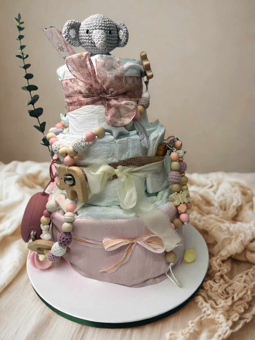 Pastel pink and mint green diaper cake baby shower centerpiece featuring three tiers of rolled white diapers wrapped in soft blush fabric and sheer ribbon bows. Top tier decorated with a crocheted gray elephant plush, accented with floral pink ribbon and delicate tulle detail. Adorned with wooden and silicone beaded pacifier clips in pastel tones, butterfly charm, wooden teething rings, and personalized letter beads spelling “BABY.” Includes pink pacifier case attached to the side. Elegant boho newborn gift