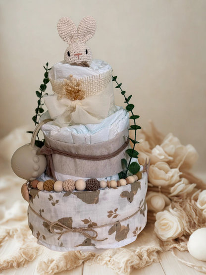 Neutral woodland bunny diaper cake baby shower centerpiece featuring three tiers of rolled white diapers wrapped in soft beige and natural linen fabrics. The base tier is covered in a delicate bunny-print cotton in warm taupe tones and tied with rustic twine for a charming countryside finish.

The middle tier is wrapped in textured oatmeal fabric and secured with natural jute string, complemented by a coordinating silicone and crochet beaded pacifier clip in shades of cream, caramel, and chocolate brown. A 