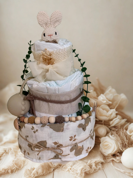 Neutral woodland bunny diaper cake baby shower centerpiece featuring three tiers of rolled white diapers wrapped in soft beige and natural linen fabrics. The base tier is covered in a delicate bunny-print cotton in warm taupe tones and tied with rustic twine for a charming countryside finish.

The middle tier is wrapped in textured oatmeal fabric and secured with natural jute string, complemented by a coordinating silicone and crochet beaded pacifier clip in shades of cream, caramel, and chocolate brown. A 