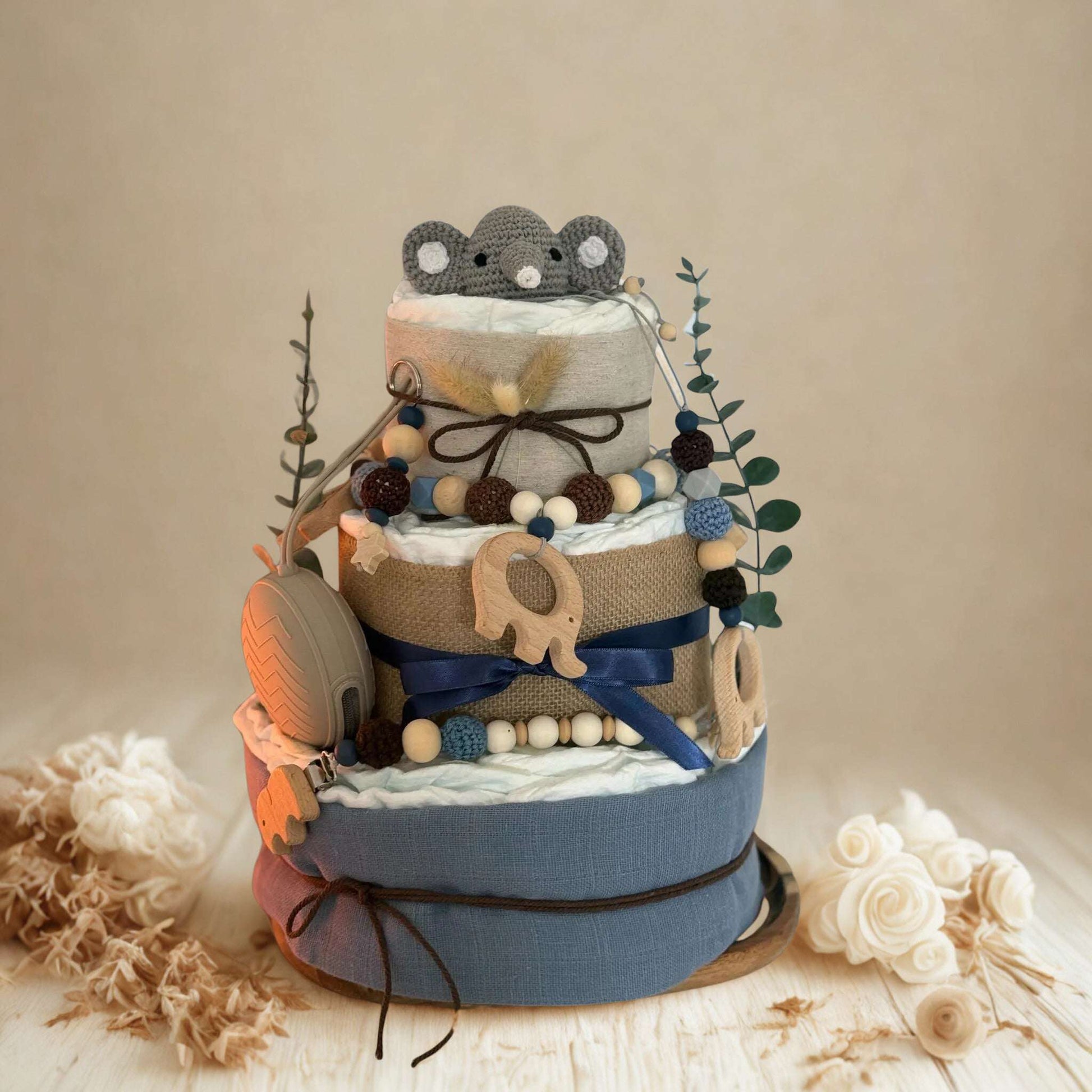 Blue and neutral safari diaper cake baby shower centerpiece featuring three tiers of rolled white diapers wrapped in soft blue fabric and natural burlap ribbon. Top tier decorated with a crocheted gray elephant plush, accented with dried pampas grass and eucalyptus greenery stems. Adorned with wooden elephant-shaped teething rings, silicone and wooden beaded pacifier clips in navy, brown, and cream tones, plus dark blue satin bow detail. Includes beige pacifier case attached to the side. Rustic boho newborn