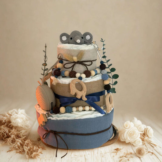 Blue and neutral safari diaper cake baby shower centerpiece featuring three tiers of rolled white diapers wrapped in soft blue fabric and natural burlap ribbon. Top tier decorated with a crocheted gray elephant plush, accented with dried pampas grass and eucalyptus greenery stems. Adorned with wooden elephant-shaped teething rings, silicone and wooden beaded pacifier clips in navy, brown, and cream tones, plus dark blue satin bow detail. Includes beige pacifier case attached to the side. Rustic boho newborn