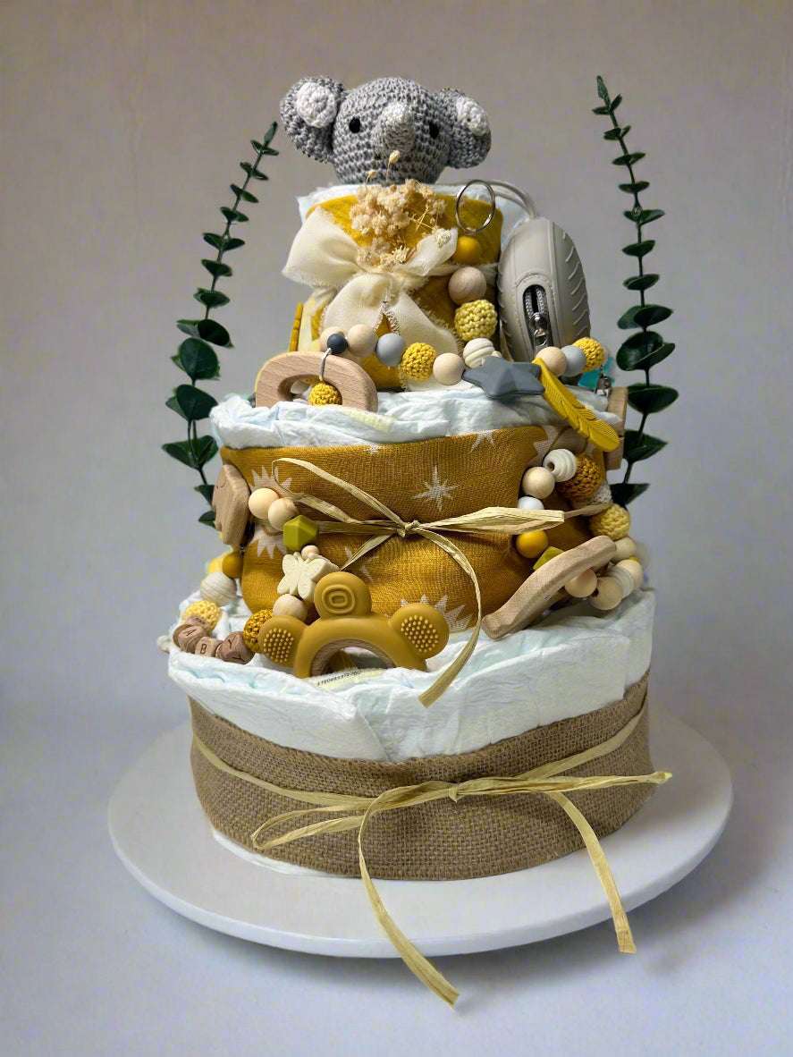 Three-tier mustard and neutral safari diaper cake featuring rolled white diapers wrapped in burlap and golden sun-print fabric, decorated with raffia bows and natural textures. A crocheted gray elephant plush sits on top, accented with dried florals and greenery. The cake includes wooden and silicone teethers, a mustard bear-shaped teether, a personalized beaded pacifier clip with wooden letter beads, and a gray pacifier case, creating a warm jungle-themed baby shower centerpiece.
