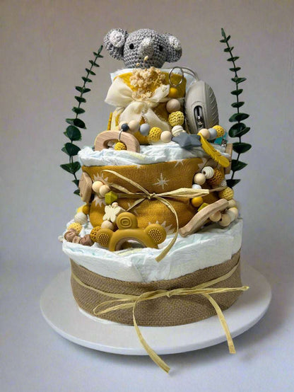 Three-tier mustard and neutral safari diaper cake featuring rolled white diapers wrapped in burlap and golden sun-print fabric, decorated with raffia bows and natural textures. A crocheted gray elephant plush sits on top, accented with dried florals and greenery. The cake includes wooden and silicone teethers, a mustard bear-shaped teether, a personalized beaded pacifier clip with wooden letter beads, and a gray pacifier case, creating a warm jungle-themed baby shower centerpiece.
