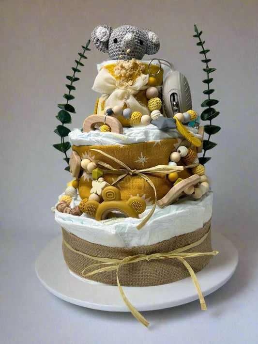 Three-tier mustard and neutral safari diaper cake featuring rolled white diapers wrapped in burlap and golden sun-print fabric, decorated with raffia bows and natural textures. A crocheted gray elephant plush sits on top, accented with dried florals and greenery. The cake includes wooden and silicone teethers, a mustard bear-shaped teether, a personalized beaded pacifier clip with wooden letter beads, and a gray pacifier case, creating a warm jungle-themed baby shower centerpiece.

