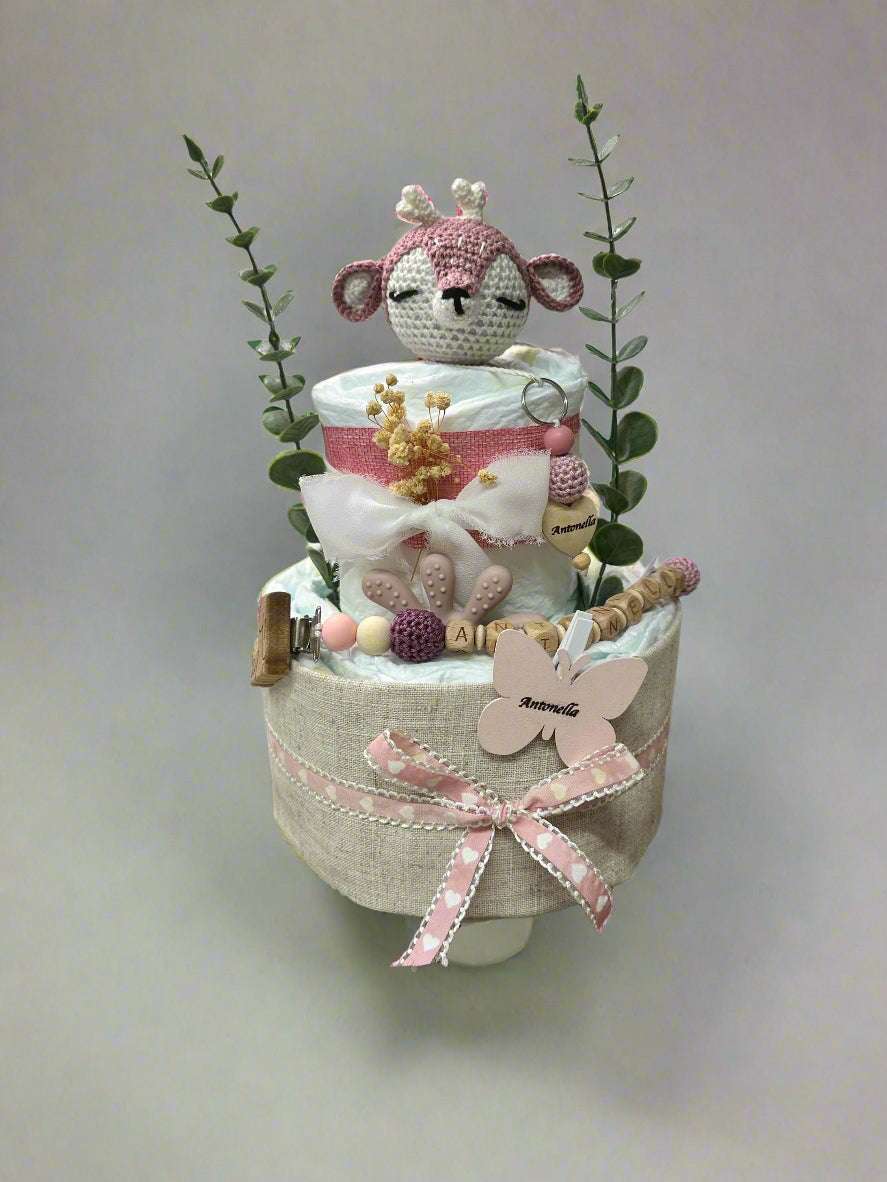 Pink and beige diaper cake baby shower centerpiece featuring two tiers of rolled white diapers wrapped in natural linen and soft pink fabric. Top tier decorated with a crocheted pink and cream deer plush toy, dried flowers, and eucalyptus greenery stems for a delicate woodland theme. Adorned with silicone and wooden beaded pacifier clip, wooden teething ring, pastel beads, and personalized name tags reading “Antonella” on a butterfly charm and wooden heart. Rustic boho newborn gift arrangement styled with p