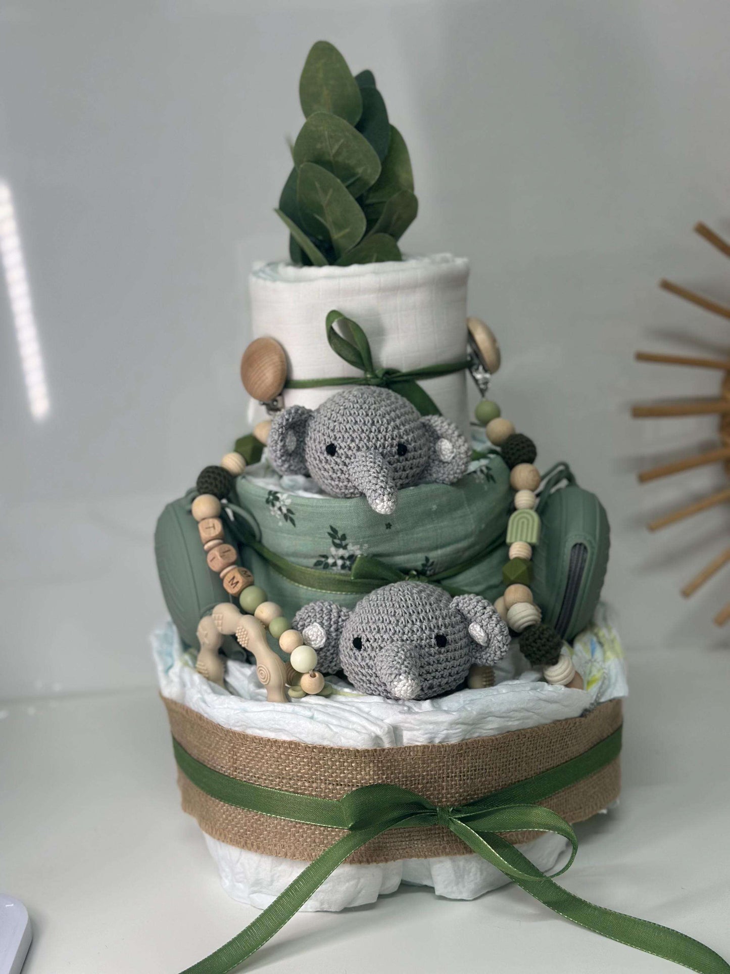 Three-tier green safari diaper cake featuring rolled white diapers wrapped in burlap and sage fabric, decorated with green ribbon bows, silicone pacifier cases, a wooden teether, and a personalized beaded pacifier clip. Two small crocheted gray elephant plush toys sit on the middle tier, topped with faux greenery, creating a neutral jungle-themed baby shower centerpiece.
