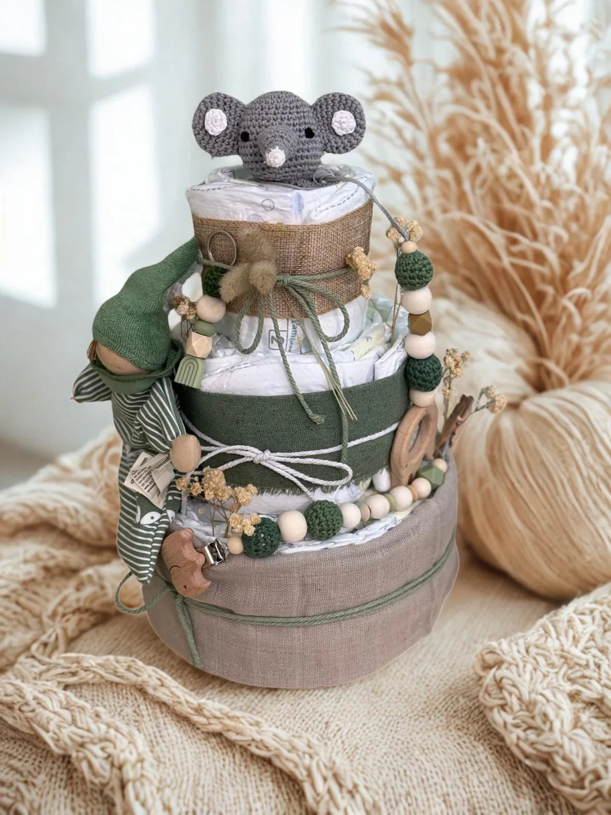 Green woodland diaper cake baby shower centerpiece featuring three tiers of rolled white diapers wrapped in sage green and natural linen fabric with rustic burlap accents. Top tier decorated with a crocheted gray elephant plush, finished with dried bunny tail grass and a twine bow for a soft boho touch. Accented with eucalyptus sprigs and delicate dried florals for a natural woodland feel.

Adorned with silicone and crochet beaded pacifier clips in deep green, cream, and natural wood tones, along with woode