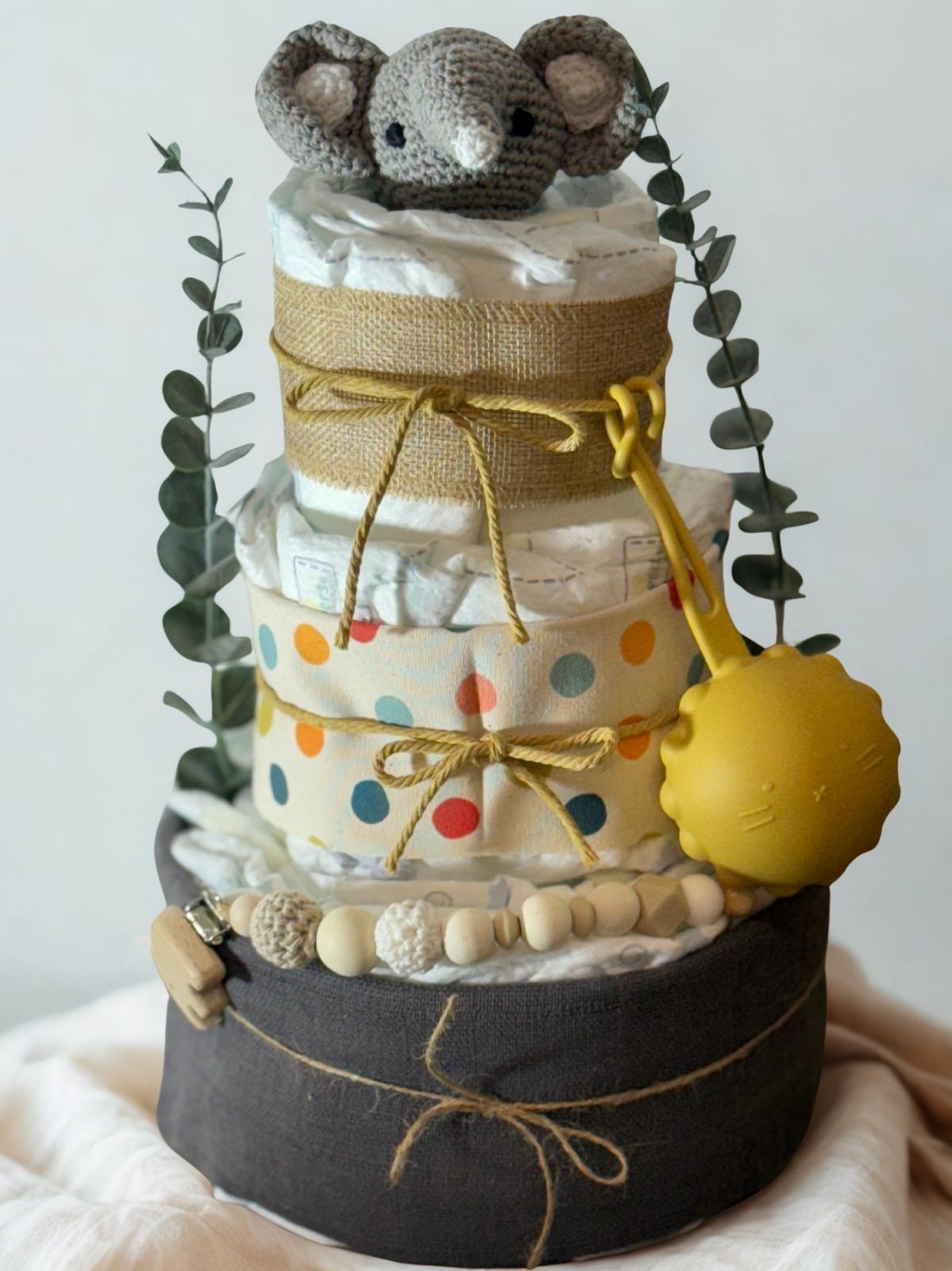 Colorful safari diaper cake baby shower centerpiece featuring three tiers of rolled white diapers wrapped in burlap ribbon and playful polka dot fabric in yellow, blue, and orange tones. Top tier decorated with a crocheted gray elephant plush, accented with eucalyptus greenery stems. Adorned with natural wooden bead pacifier clip, crochet beads in cream and beige shades, and rustic twine bows. Includes bright yellow pacifier case attached to the side for a cheerful pop of color. Modern boho newborn gift arr