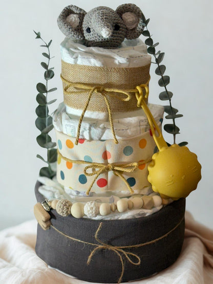Colorful safari diaper cake baby shower centerpiece featuring three tiers of rolled white diapers wrapped in burlap ribbon and playful polka dot fabric in yellow, blue, and orange tones. Top tier decorated with a crocheted gray elephant plush, accented with eucalyptus greenery stems. Adorned with natural wooden bead pacifier clip, crochet beads in cream and beige shades, and rustic twine bows. Includes bright yellow pacifier case attached to the side for a cheerful pop of color. Modern boho newborn gift arr