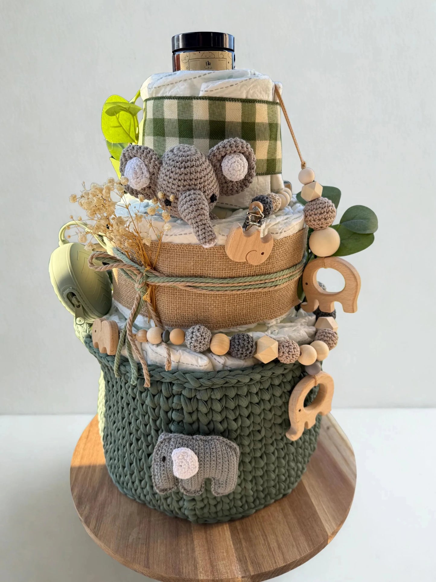Green safari diaper cake baby shower centerpiece featuring three tiers of rolled white diapers wrapped in burlap ribbon and green gingham fabric. Top tier decorated with a crocheted gray elephant plush, with a baby care jar placed above. Accented with dried baby’s breath flowers, eucalyptus leaves, and rustic twine bows for a natural woodland theme. Adorned with wooden elephant-shaped teething rings, silicone and crochet beaded pacifier clip in sage, beige, and gray tones. Includes matching green pacifier c